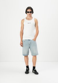 Hvit ermeløs tanktop med svart tekst, lyseblå denimshorts, svarte sneakers med hvite striper, og svarte solbriller.