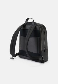 Mochila preta com uma mistura de materiais lisos e texturizados, apresentando alças de malha almofadadas, zíperes duplos e uma forma retangular e aerodinâmica.