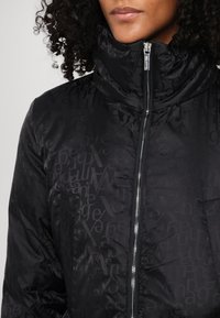 Black puffy jacket with high collar, featuring a subtle tonal pattern of letters. Zipper closure with silver hardware and side pockets.