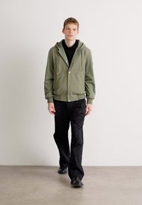 Chaqueta verde claro con capucha y botones plateados, suéter negro de cuello alto debajo y pantalones negros holgados. Modelo camina sobre un suelo gris.
