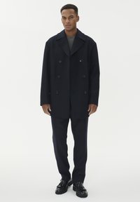 Cappotto in lana navy a doppio petto con grandi bottoni, abbinato a un maglione grigio e pantaloni scuri, indossato con scarpe nere.