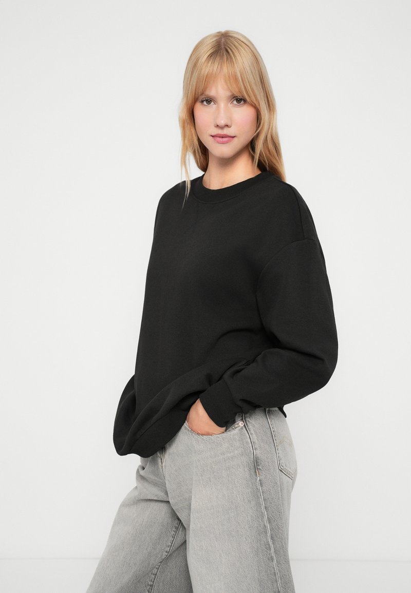 Jonge vrouw met blond haar die een zwart oversized sweatshirt en lichtgrijze jeans draagt, staande met een hand in haar zak.