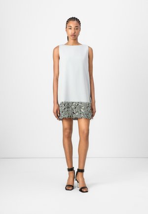Robe sans manches de couleur gris clair avec un ourlet texturé et frangé de couleur grise. Le mannequin porte des sandales à talons noires, se tenant devant un fond blanc uni.