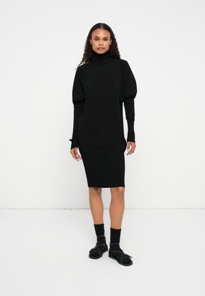 Rochie neagră cu model în relief, cu guler tunică și mâneci bufante, până la genunchi, cu manșete ajustate. Este purtată cu șosete negre și sandale.