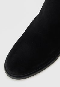 Stivaletto in suede nero con punta arrotondata, texture liscia e una sottile bordatura in materiale contrastante.