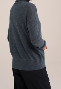 Pull en maille gris foncé avec un col montant, une coupe ample et des poignets côtelés. Texture douce, design minimaliste et couleur unie.