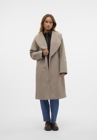 Cappotto beige con colletto drappeggiato, maniche lunghe e chiusura frontale. Indossato su un top nero e jeans blu, abbinato a scarpe marroni scure.
