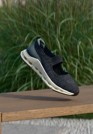 Schwarzer gestrickter Mary Jane Sneaker mit weißer, gepolsterter Sohle, schwebt über einer Holzbank mit grünem Laub im Hintergrund.