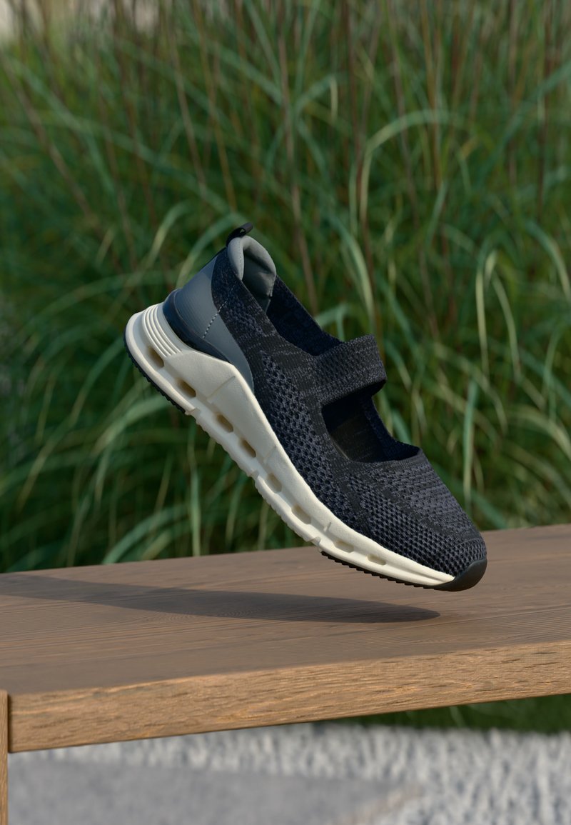 Μαύρο πλεκτό αθλητικό παπούτσι Mary Jane με λευκή μαλακή σόλα, που αιωρείται πάνω από ένα ξύλινο πάγκο με πράσινα φύλλα στο παρασκήνιο.