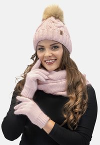 Berretto lavorato a maglia rosa con pompon, sciarpa rosa e guanti. La texture è morbida, con un motivo a costine sul berretto e sui guanti.