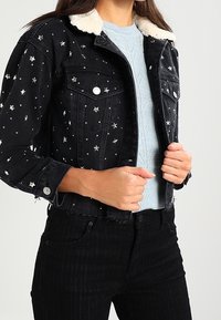 Černá džínsová bunda se stříbrnou výšivkou hvězdy, límcem z umělé kožešiny, zapínáním na knoflíky a dvěma náprsními kapsami, v kombinaci s světlemodrým svetrem.