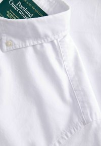 Bijela košulja s dugim rukavima i strukturiranim ovratnikom, izrađena od glatkog pamuka. Na oznaci piše "Portland Outerwear", prikazujući logo i naziv brenda.
