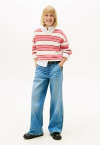Maglione a righe rosse e bianche con collo rotondo, abbinato a jeans a gamba larga blu chiaro e scarpe nere, in piedi su uno sfondo bianco.