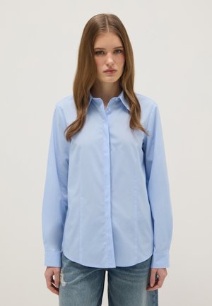 Mujer joven con cabello largo y castaño, vistiendo una camisa azul claro abotonada y jeans azules desgarrados sobre un fondo liso.