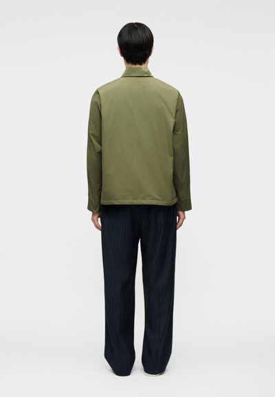 Άνδρας, φαίνεται από πίσω, φοράει ένα μπουφάν με κουμπιά ελιάς και άνετο σκούρο μπλε παντελόνι με ρίγες πάνω σε απλό φόντο.