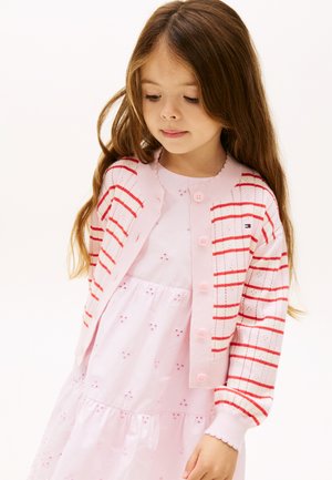 Noor tüdruk, kellel on pikad pruunid juuksed, kannab heleroosat kleiti ja punaste triipudega roosat kardigani, vaadates vasakule alla.