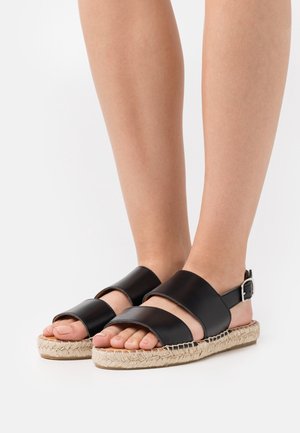 Jalat mengenakan sandal kulit hitam dengan dua tali lebar dan sol datar yang dibungkus jute, di latar belakang putih.