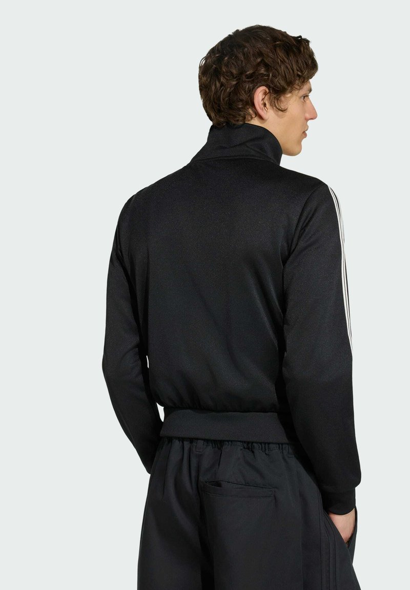 Junger Mann mit lockigem braunem Haar, der eine schwarze Trainingsjacke mit weißen Streifen an den Ärmeln und passende schwarze Hose trägt, von hinten.