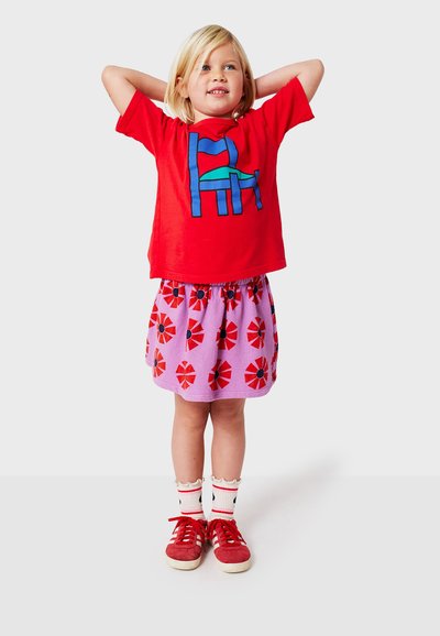 Rød kortærmet t-shirt med blå og grøn grafisk motiv, parret med en lyserød nederdel med røde blomstermønstre. Afsluttet med røde sneakers.