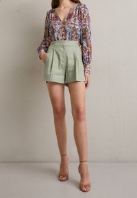 Bloemenblouse met lange mouwen en V-hals, gecombineerd met lichtgroene shorts met hoge taille, plooien en zijzakken. Huidskleurige hakken.