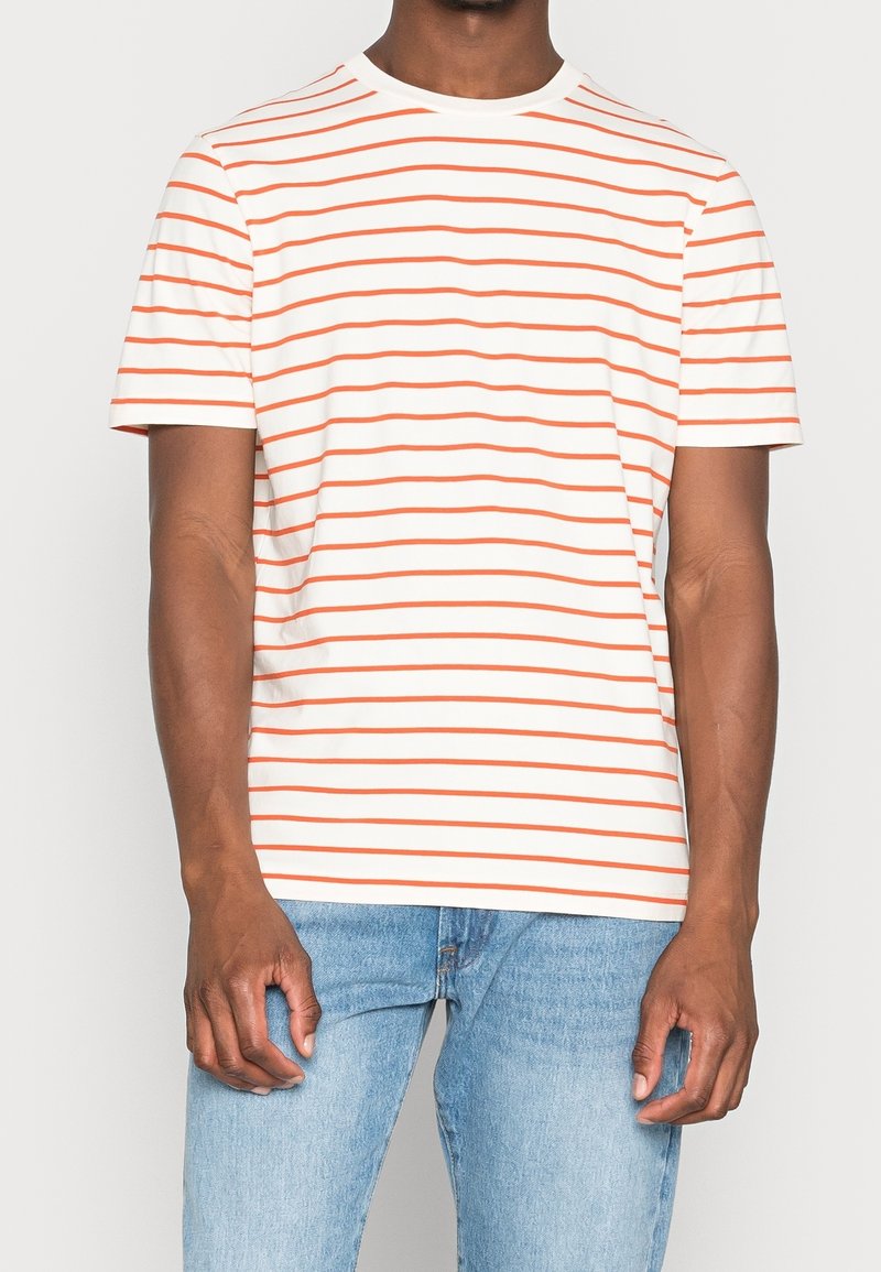 Hombre vestido con una camiseta de manga corta blanca con finas rayas horizontales naranjas y jeans de color azul claro, de pie con los brazos relajados.
