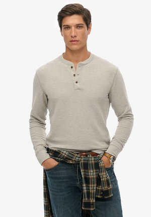 Helles graues Langarm-Henley-Shirt mit drei braunen Knöpfen, kombiniert mit dunklen Jeans und einem karierten Hemd, das um die Taille gebunden ist.