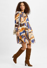 Chiffonkleid mit einem abstrakten Muster in Blau, Orange und Braun, langen Ärmeln, runder Halslinie und lockerem Schnitt. Braune Stiefel bis zum Knie.