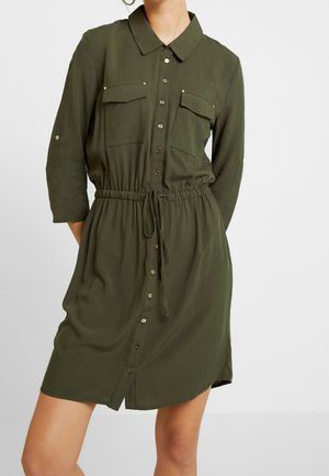 Nainen, joka pukeutunut oliivinvihreään nappitolppaiseen paitamekkoon, jossa on kiristysnauha vyötäröllä ja kaksi rintataskua, seisoo yksiväristä taustaa vasten.