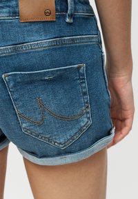 Τζιν σορτς με γυρισμένα μανίκια, πίσω τσέπη με πορτοκαλί ραφές, δερμάτινο σήμα μάρκας, φορεμένα από πρόσωπο με ορατό χέρι στο πλάι.