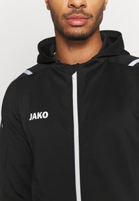 Zwarte zip-up jack met capuchon, witte rits en het "JAKO" logo in het wit op de linkerborst. Getextureerde stof met contrasterende schouderaccenten.