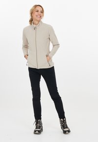 Fleecejakke i lys beige med høj krave, lynl�ås fortil og sidelommer. Båret med sorte bukser og grå vandrestøvler.