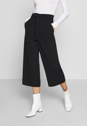 Pantalons larges noirs à taille haute avec détail de ceinture, featuring des poches latérales et une texture de tissu lisse. Associé à des bottines blanches.