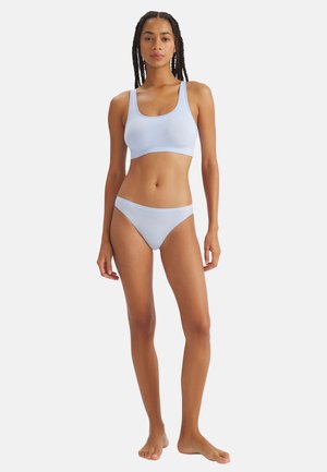 Frau mit langen Zöpfen steht barfuß und trägt ein hellblaues Sport-BH und passende Bikini-Unterwäsche vor weißem Hintergrund.