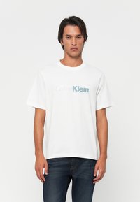 Mand iført en hvid Calvin Klein New York T-shirt og mørke jeans, stående mod en simpel hvid baggrund.