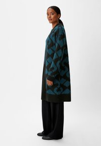 Langer Cardigan in Dunkelgrün und Petrol mit geometrischen Mustern. Hergestellt aus einem weichen Material, mit lockerer Passform und Taschen, kombiniert mit schwarzen Hosen.