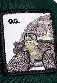 Izšūta nozīmīte ar bruņurupuci uz baltas fona ar melnu malu. Teksts "O.G." ir melnā krāsā augšā. Zaļas auduma pamatne.