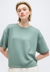 Νεαρή γυναίκα με κοντά ξανθά μαλλιά που φοράει φαρδιά πράσινη πουκαμίσα με κοντά μανίκια και λευκή φούστα, στέκεται μπροστά σε απλό φόντο.