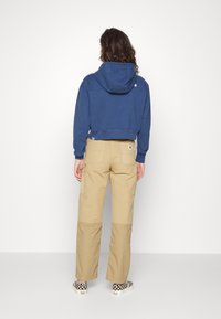 Felpa blu navy corta con cappuccio, pantaloni cargo beige con tasche e scarpe slip-on a scacchi bianchi e neri.