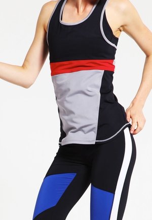 Mujer con camiseta deportiva sin mangas en negro, gris y rojo con leggings negros con paneles azules y blancos, posando con una mano en la cadera.