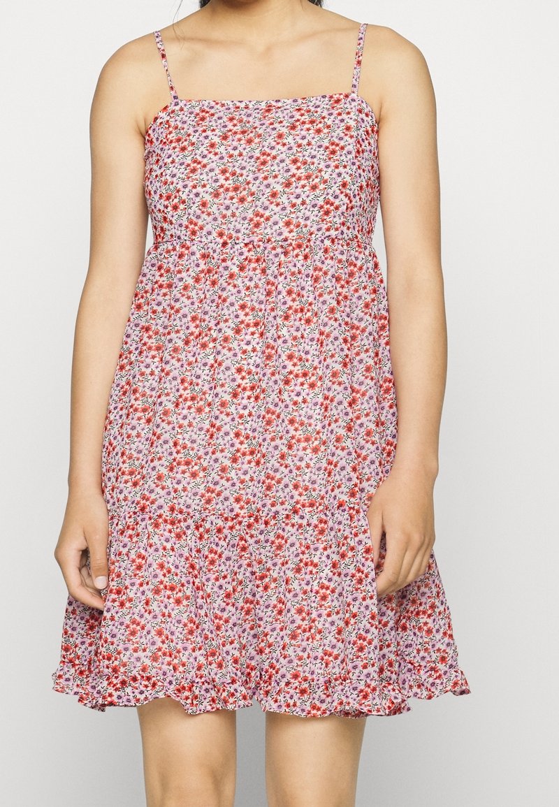 Femme portant une robe rose sans manches à bretelles fines avec un motif floral rouge, se tenant devant un fond blanc uni.