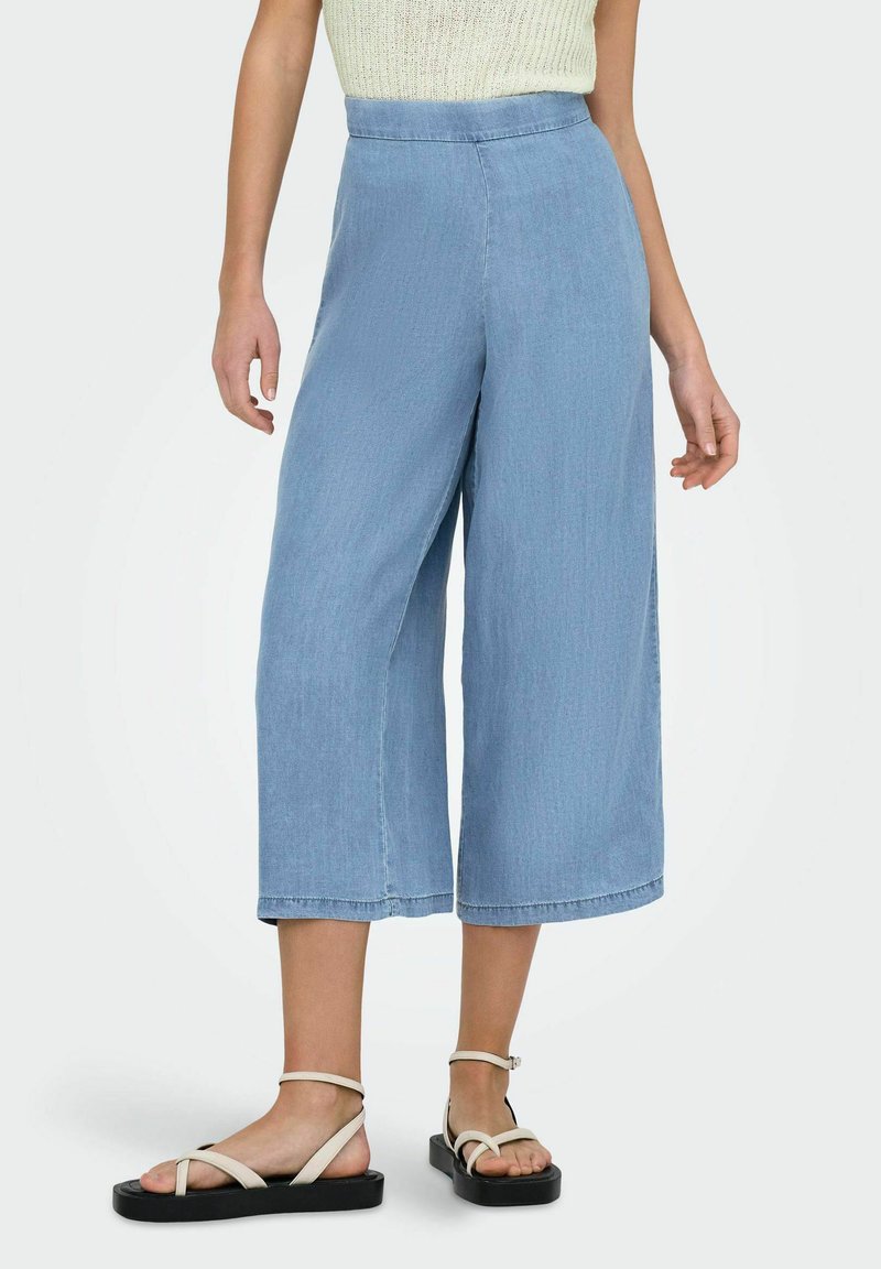 Pantalones de pierna ancha en azul claro con una cintura alta, hechos de denim suave. El dobladillo tiene un acabado de borde sin acabar, que complementa unas sandalias de tiras beige.
