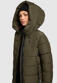Olivenfarvet polstret jakke med en overdimensioneret hætte, lynlåslukning og sort hardware. Har en quiltet tekstur og sidelommer.