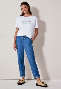 T-shirt blanc à manches courtes avec imprimé "Breeze", associé à un pantalon en denim décontracté bleu clair et des sandales noires avec un dessus en paille tressée.