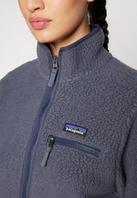 Giacca in pile di un elegante blu navy con colletto alto, zip integrale e tasca sul petto. Presenta una superficie testurizzata e un logo Patagonia.