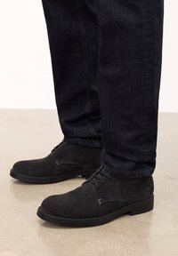 Botas de tobillo de ante negro con diseño de cordones, punta redondeada y suela de goma baja. Combinadas con unos jeans de mezclilla oscuros, mostrando un primer plano de los zapatos.