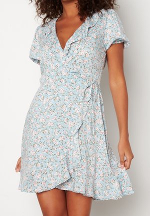 Woman wearing a light blue floral wrap dress with ruffle trim and short puff sleeves, holding the hem slightly lifted.