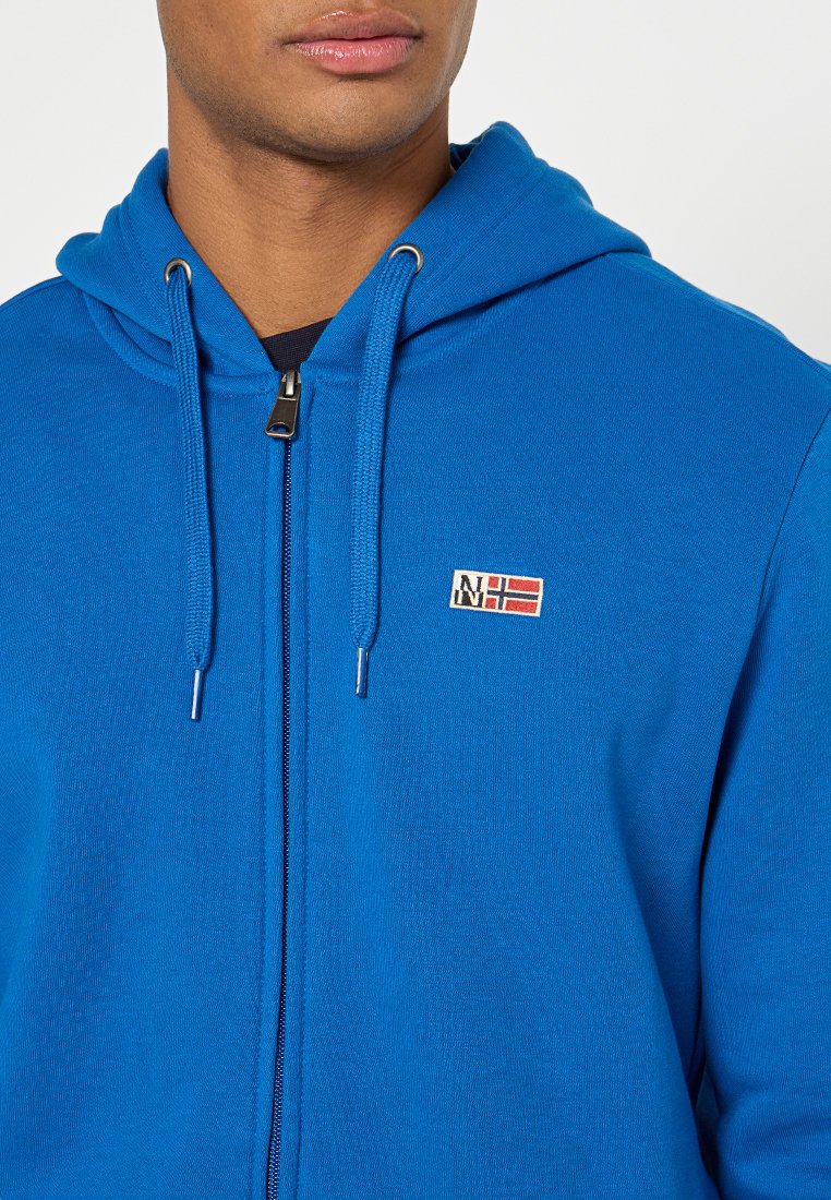 Hoodie zippé bleu vif avec cordons de serrage, présentant un petit patch brodé avec des accents rouges et blancs. Tissu en tricot doux et coupe standard.