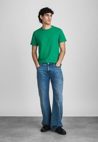 Fiatal férfi zöld pólót, kék farmert és fekete cipőt visel, a zsebébe dugott kezével áll, a háttér előtt oldalt néz.