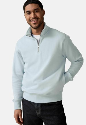 Felpa con zip blu chiaro in tessuto morbido, con colletto alto e polsini a costine, indossata con jeans scuri.