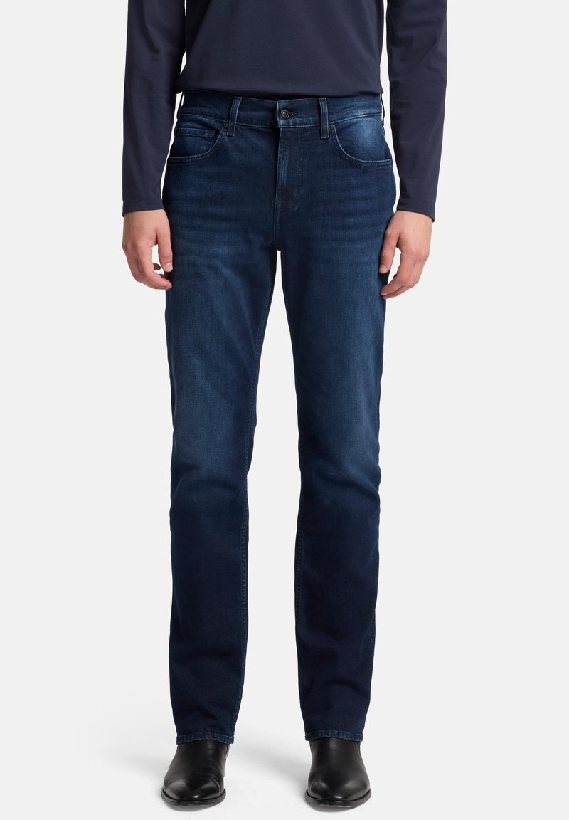 Jeans in denim blu scuro con taglio a gamba dritta, caratterizzati da cinque tasche e cuciture bianche a contrasto, abbinati a stivali in pelle nera.